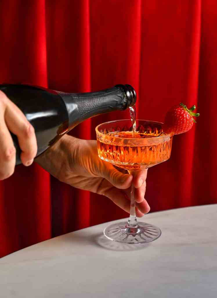 A hand pouring a wine into a coupe glass on the red curtain background